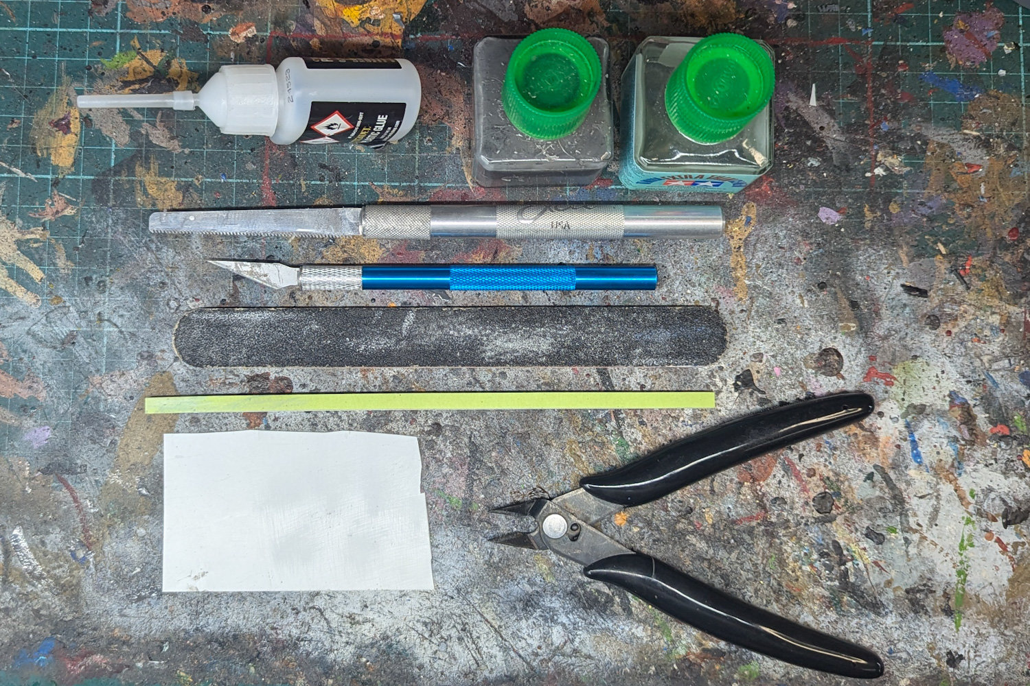 Image depicts the nine following tools, Hobby Knife,  Hobby Saw,  Clippers,  Plastic Glue,  0.5mm plasticard sheet, Foam Nail File,  Sanding Sticks,  Tamiya Thin Glue,  Sprue Goo